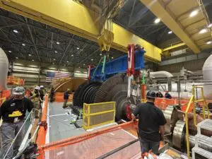 Industrial facility with a large yellow overhead crane, heavy machinery, and workers in hard hats and vests examining equipment.