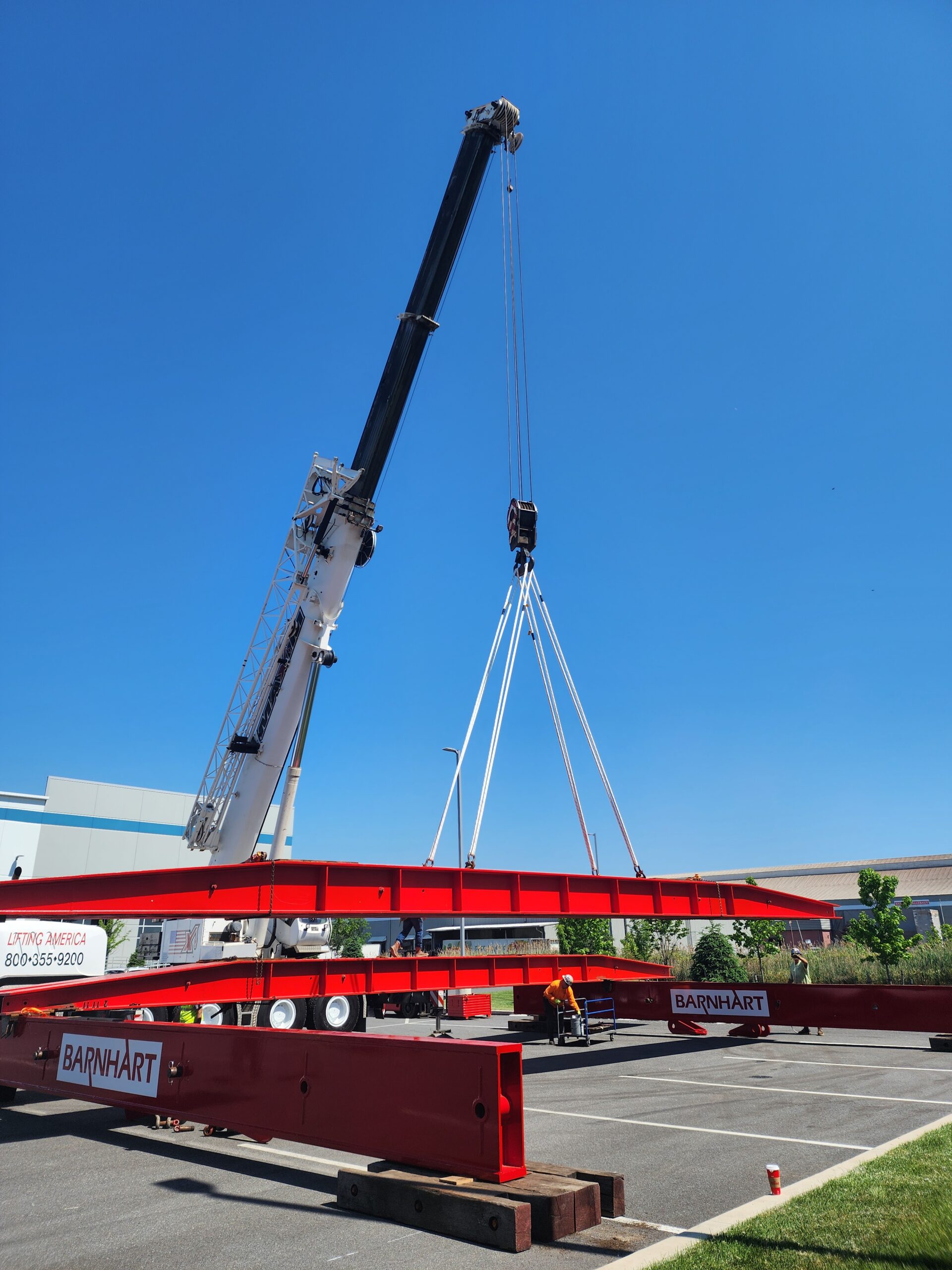 20240522_104208 | Slingmax® Rigging Solutions | Official Site | Twin-Path® Slings Crane lifting a red steel beam across a parking lot under a clear blue sky.